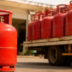 A red commercial LPG cylinder placed in the foreground with multiple LPG cylinders loaded on a delivery truck outside a gas distribution facility, representing commercial LPG supply and pricing.