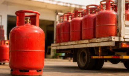 A red commercial LPG cylinder placed in the foreground with multiple LPG cylinders loaded on a delivery truck outside a gas distribution facility, representing commercial LPG supply and pricing.